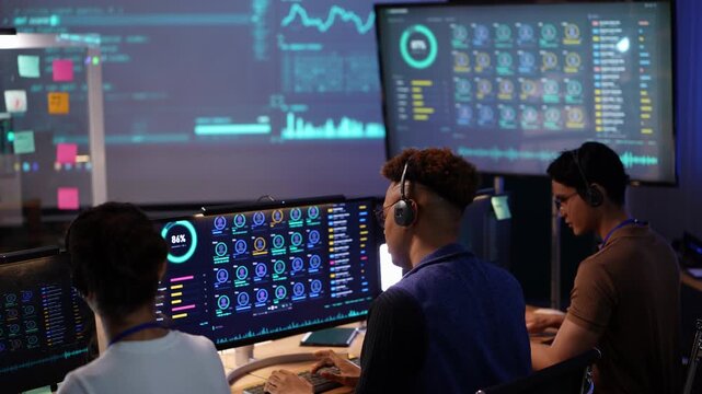 Rear view of three operators at a dashboard workstation, an Asian man in a brown polo adjusts his headset and stands up while teammates keep typing under the data wall
