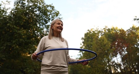 Senior woman exercising with hula hoop outdoors, low angle view. Space for text © New Africa