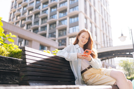Happy young woman with long red hair sitting on bench in urban setting. Redhead female smiling using mobile phone on bright, sunny day. Modern city building in background. Casual lifestyle concept.