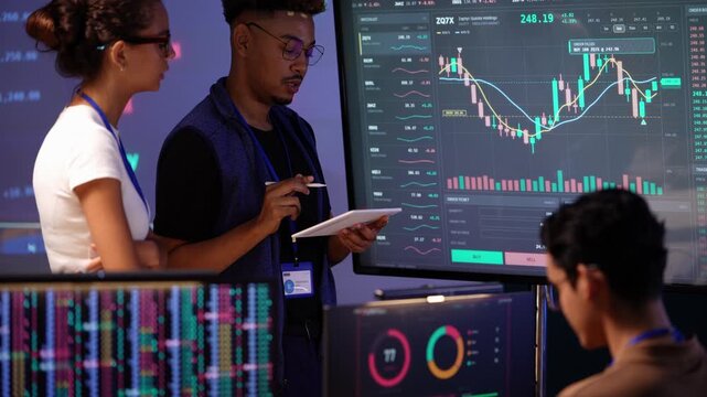Latina woman gestures and smiles as she talks with a Black colleague holding a tablet and stylus beside a live stock chart, an Asian teammate seated in the foreground