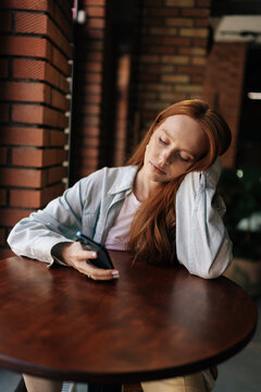 Exhausted young woman with long red hair rests head on hand at cafe table. Sleepy female with closed eyes holding smartphone, feeling burnout, boredom in cozy restaurant with warm lighting.