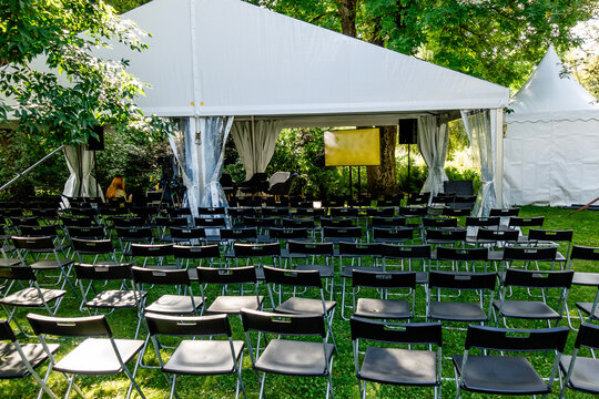 Outdoor event setup in city park with white canopy and rows of black chairs prepared for conference, lecture, discussion, or public event