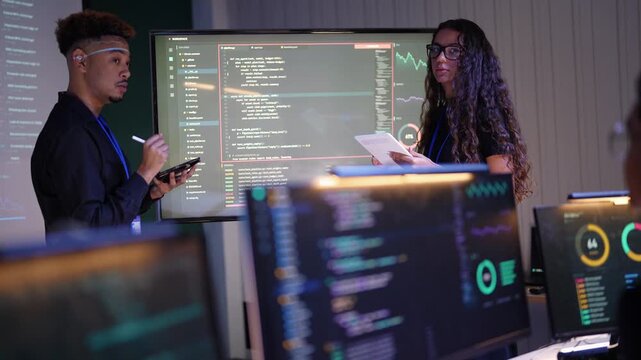 Black developer writes on his tablet while a Latina colleague with long curly hair and glasses holds a printout and talks beside a huge code screen, a blurred coworker at the edge