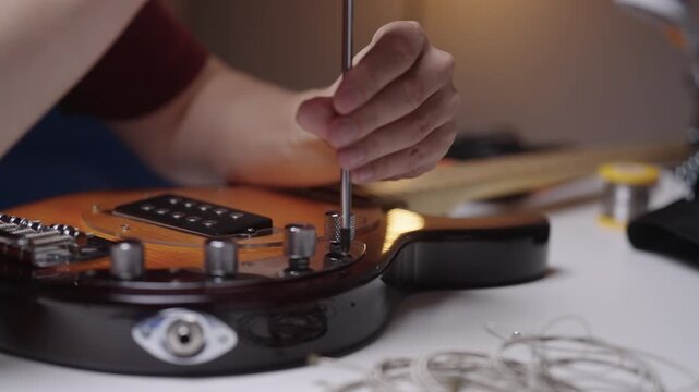 Skilled luthier hands performing professional maintenance on sunburst electric guitar, installing new strings and adjusting tuning pegs with precision tool in workshop, close-up, slow motion.