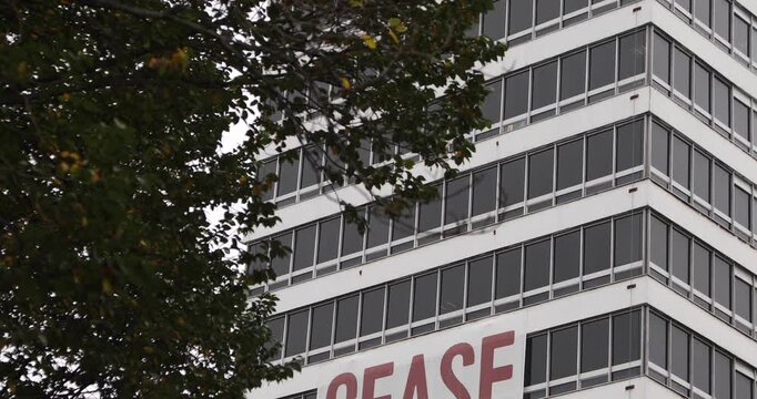 A large &ldquo;CEASE FIRE NOW&rdquo; banner hangs on an urban office building, framed by a tree, expressing an urgent call for peace, civilian protection and an end to armed conflict.