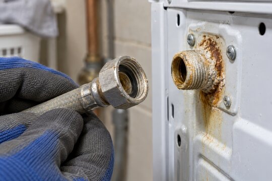 Gloved hand disconnecting a corroded washing machine hose with rusty, scaled pipe fitting. Appliance plumbing repair and maintenance. Hard water buildup and household wear.