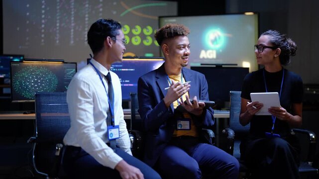 Black engineer laughs and talks with his hands as the Asian colleague gestures back and the Latina engineer smiles, a large AGI logo glowing on the display behind the seated team