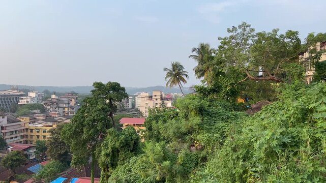 Panning shot of Panaji city skyline from Altinho hill showing urban buildings, palm trees and Mandovi river in Goa, India