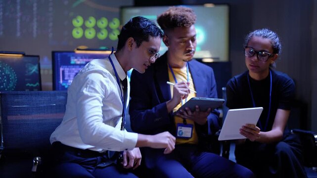 Asian developer gestures above his head, then he and a Latina coworker lean in to look at the tablet a Black teammate holds, the three huddling over the screen in a blue lit lab