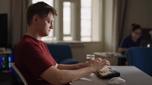 Side view of casual young man in profile working on computer, typing on split keyboard, taking break to drink from glass, with coworker sitting at another desk in background. Shooting in slow motion.