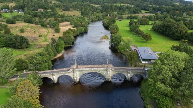 Aerial footage of the historic Wade's Bridge over the River Tay in Aberfeldy, Perthshire, Scotland. A single car crosses the stone bridge, surrounded by green countryside, trees and a golf course