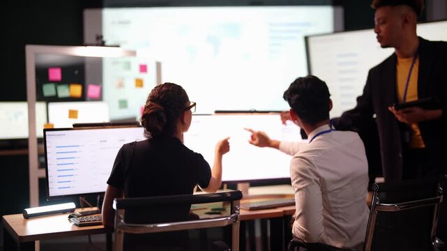 Black team lead stands with a tablet and pen watching as a Latina developer and an Asian coworker seated with their backs to camera both point at a chart on a bright monitor