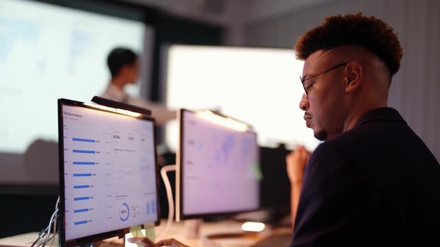 Black engineer looks down at his dashboard then turns toward an Asian presenter at the big screen, a Latina coworker with her hair in a bun sits beside him, layered focus