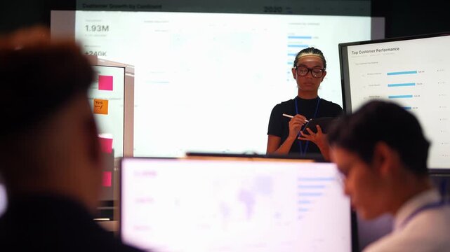 Latina woman in glasses turns from the projected world map, faces the room holding a tablet, then points her stylus at a performance bar chart as Asian and Black coworkers listen.