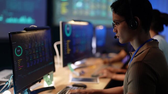 Focused Asian developer in glasses and headset looks down and types on his keyboard, glances at the dashboard monitor, then types again as a Latina colleague works at the next desk.