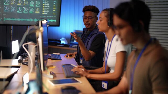 Black man with blond curly top scratches his head unsure, gestures while thinking aloud, then covers his mouth in thought as a Latina teammate types and an Asian man sits blurred.