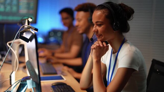 Smiling Latina woman with a headset talks on a call, rests her chin on clasped hands and laughs, while a Black and an Asian colleague type behind her at a long row of desks.
