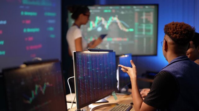 Latina woman holding a tablet explains a candlestick chart on the big screen as a Black programmer at his desk stops typing, clasps his hands and listens with an Asian coworker