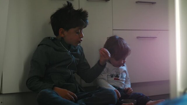 Caucasian brothers sitting on the kitchen floor sharing a snack
