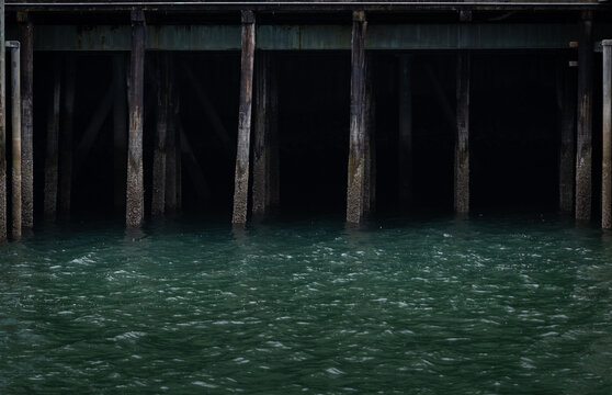 Photo of darkness under a high pier large supporting pillars calm water. Marina abstract background