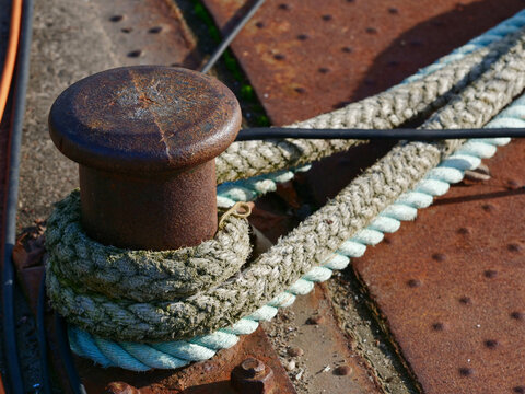 A rusty cast-iron mooring bollard with weathered mooring ropes wrapped around it on a textured, rusty metal ground.