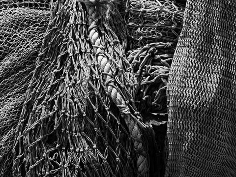 A high-contrast black and white close-up view of tangled commercial fishing nets with various mesh sizes, highlighting a thick braided rope and intricate textures.