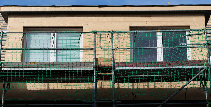 a building under construction, showing a newly installed window set into light wood cladding, protected by construction scaffolding with a green safety net.