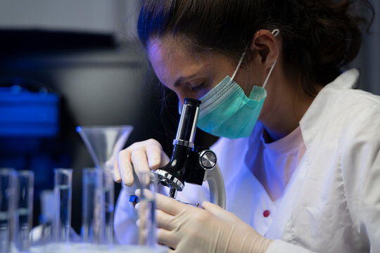 Professional female biologist in protective gear analyzing microbiology sample under lab microscope lens.