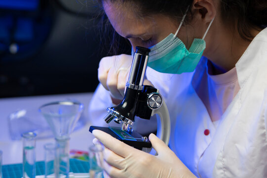 Female researcher in sterile mask placing glass sample slide onto microscope stage for scientific study.