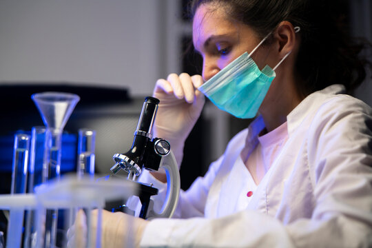 Tired female scientist adjusting medical face mask during long microscope research in dark laboratory.