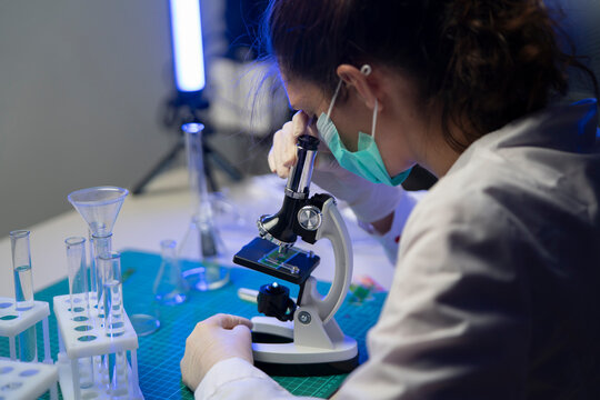 Medical researcher sitting at desk looking through microscope during clinical study in neon lit laboratory.