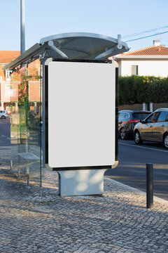 Blank outdoor advertising panel installed at a modern glass bus shelter on a residential street in Portugal. Parked cars, tiled pavement, nearby houses and a clear blue sky create a typical urban sett