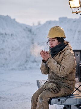 Young female construction worker in yellow hard hat warming hands with breath while sitting on truck tailgate in freezing snowy environment