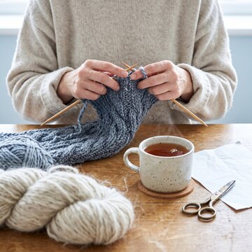 Unrecognizable woman in a cozy sweater knitting a chunky grey wool scarf with wooden needles at a table alongside a hot cup of tea and yarn