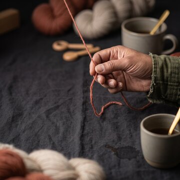 Artisan hand holding terracotta yarn strand over dark linen table with ceramic coffee mugs and twisted wool skeins in cozy studio