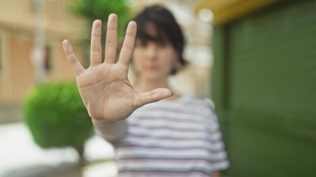 Young woman holds open palm toward camera on a street beside a green shutter and trimmed bush; boundary defiance.