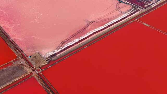 Geometric pattern of red salt evaporation ponds divided by railway tracks. Aerial top down shot displays vivid pink rectangular mineral basins separated by dirt roads and narrow transport lines.
