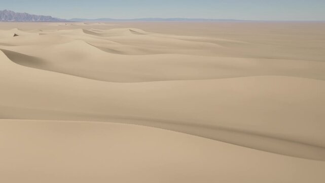 A wide view shows vast sand dunes in a desert area. The dunes vary in shape and size. The landscape has no vegetation and appears dry and warm. The sky is clear and blue, hinting at daytime.