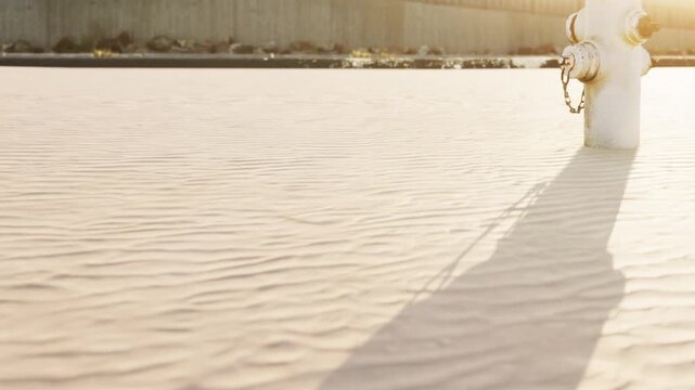A fire hydrant stands on sandy ground as the sun sets, creating a long shadow. The scene shows a simple urban landscape with a wall in the background.