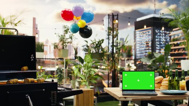 Green screen laptop mockup sits beside drinks and bread on a rooftop barbecue table, providing an editable display for outdoor event advertising.