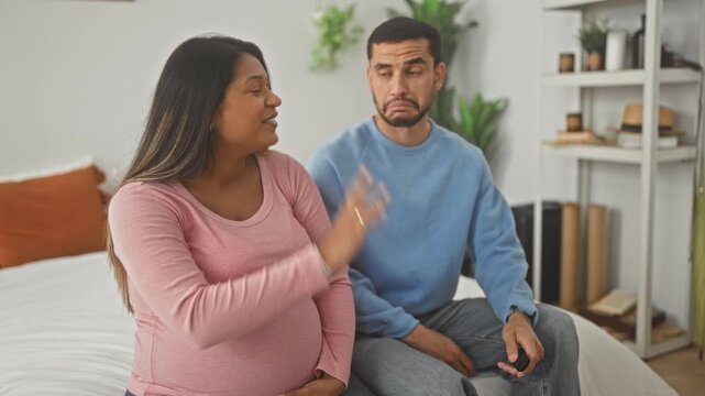 Pregnant woman with hand on belly gestures to man who holds a tv remote while they sit on a bed in a building; expecting parents concern.