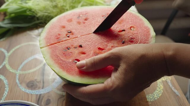Hands slice watermelon with care and precision. Red flesh and green rind reveal sweet, juicy interior. Fresh fruit sits on wooden board beside lettuce. Sunlight highlights the vibrant colors