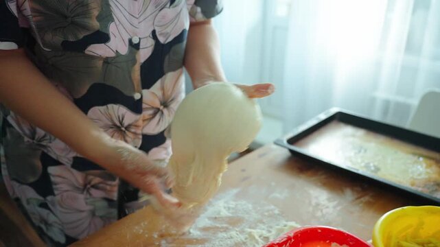 Hands knead dough with flour on wooden surface. Dough is soft, round, and ready for shaping. Warm kitchen light shows cozy, homey activity. Cooking process feels personal and comforting