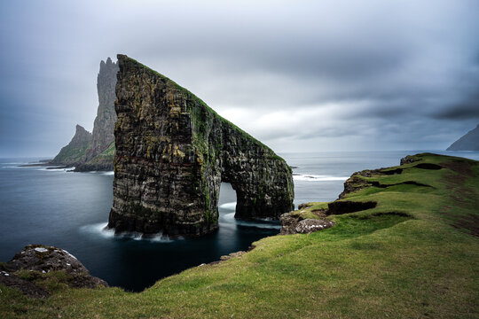 View of the Drangarnir sea stacks rising from the ocean with dramatic cliffs and lush green grassy slopes under a cloudy sky in the Faroe Islands.