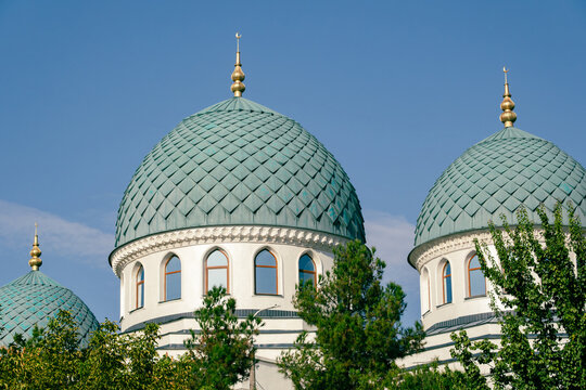 View of the green tiled domes and golden finials of the Minor Mosque surrounded by trees under a clear blue sky in Tashkent, Uzbekistan.