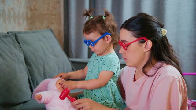 Mother and child play with dolls at home. They wear colorful glasses, smiling while engaging. Soft couch and curtains frame their cozy moment. Ideal for parenting, family, or playful domestic scenes