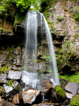 View of Melincourt Falls cascading over rocky cliffs surrounded by lush green foliage and mossy rocks in Neath, Wales, United Kingdom.