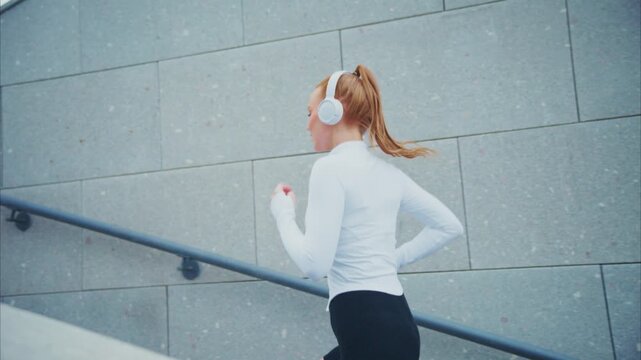 Young woman runs up stairs with headphones on. Her hair bounces as she moves steadily upward. Grey concrete walls frame her athletic stride. She appears focused, undeterred by the incline