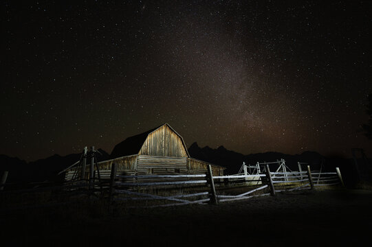 View of the historic wooden barn at Mormon Row under a starry night sky with the Milky Way galaxy visible above the Grand Teton mountains in Teton County, Wyoming, United States.