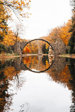 View of The Devil's Bridge arching over a calm pond surrounded by vibrant autumn foliage in the forest of Gablenz, Sachsen, Germany.